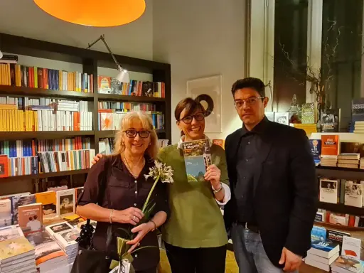 Tre persone sorridono in una libreria, una tiene in mano il libro "Cocco e Magella". Libri in ordine cronologico sugli scaffali.
