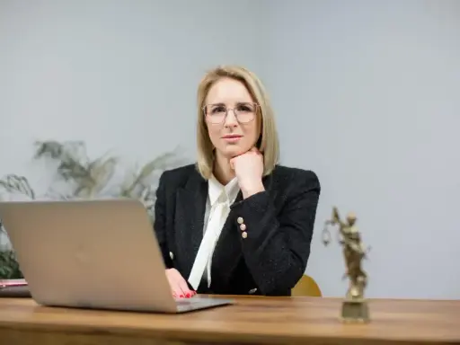 Kobieta w okularach i żakiecie siedzi przy biurku z laptopem i statuetką Temidy.