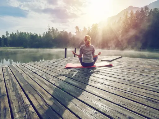 En kvinna med ryggen mot kameran sitter i meditationsställning på en brygga vid en sjö. Solen strålar genom dimman och skapar en magisk stämning. Hon verkar vara helt i nuet.