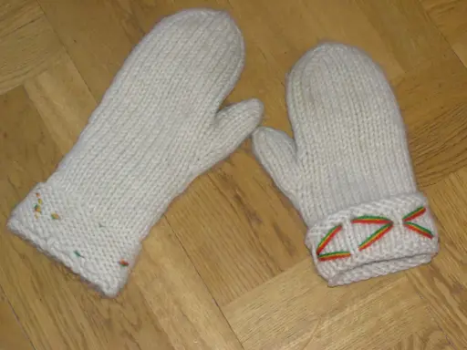 Två vita, stickade vantar med diskreta drops design-mönster i färggranna trådar på muddarna.