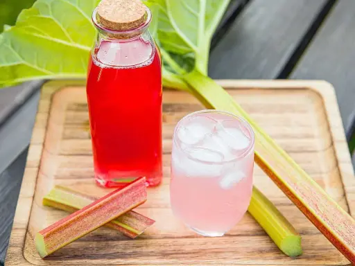 En flaska med röd rabarbersaft och ett glas med is och rosa rabarbersaft står på en träbricka. Rabarberstjälkar ligger bredvid.