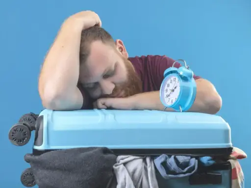 Zmęczony podróżnik śpi na walizce, budzik obok sygnalizuje jetlag.