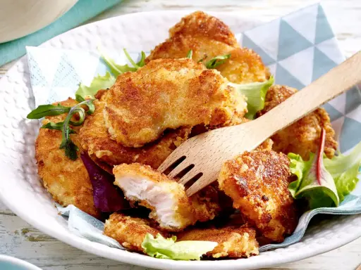 Knusprige Hähnchen-Nuggets, perfekt im Butterschmalz gebraten, auf einem Bett aus Salat. Ein Holzgabel liegt bereit.