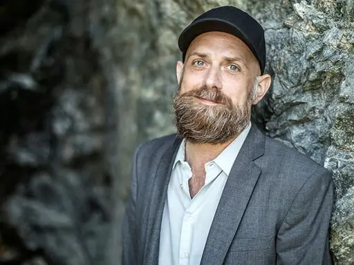 Man med skägg och keps, i grå kavaj, står mot en stenvägg. Kanske en del av en förintelsen serie på SVT.