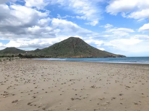 Weite Sandfläche am Playa de los Genoveses mit sanften Wellen und einem grünen Berg im Hintergrund unter einem bewölkten Himmel.
