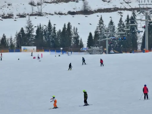 Narciarze i snowboardziści szusują po ośnieżonym stoku w Bukowinie Tatrzańskiej. W tle widać wyciąg krzesełkowy i las.