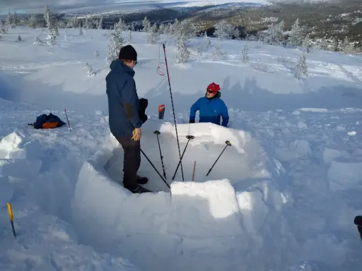 Två personer bygger en bivack i snön. De använder snöblock för att bygga väggarna och stavar för att markera utrymmet.