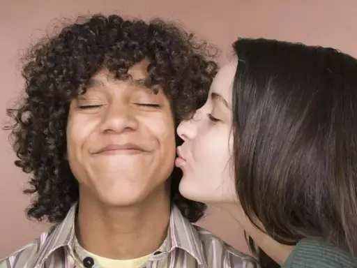 Un jeune homme aux cheveux bouclés reçoit un baiser sur la joue d'une jeune femme. Il semble ravi, les yeux fermés. Un moment de tendresse, un vrai pécho def.