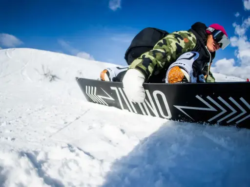 Snowboardzista w kamizelce moro i różowej czapce, zjazd na desce. Warto wiedzieć, jak dobrać buty snowboardowe, by czuć się pewnie na stoku.