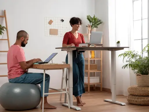 Un homme travaille sur son ordinateur, assis sur un ballon de gym, tandis qu'une femme travaille debout à un bureau réglable. Un espace de travail moderne et dynamique.