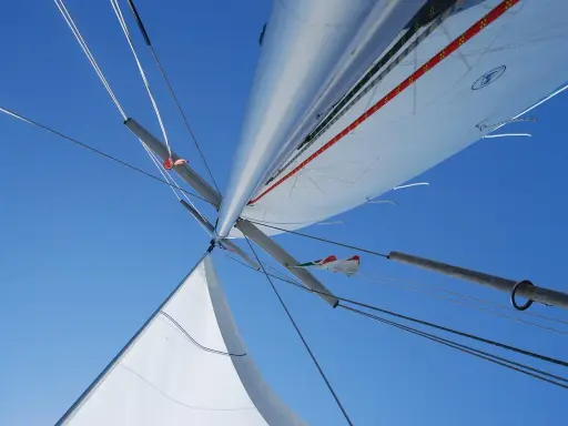 Vue plongeante sur le mat de bateau, voiles blanches déployées sous un ciel bleu éclatant, cordages tendus.