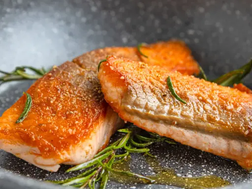 Zwei perfekt gebratene Lachsfilets mit knuspriger Haut und Rosmarin in einer Pfanne.