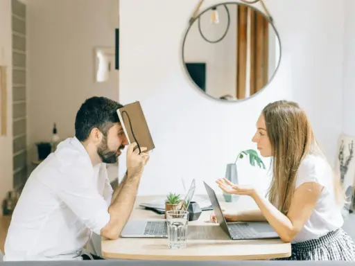 Hombre con personalidad narcisista, frustrado, se cubre la cara con un libro mientras una mujer le habla con exasperación.
