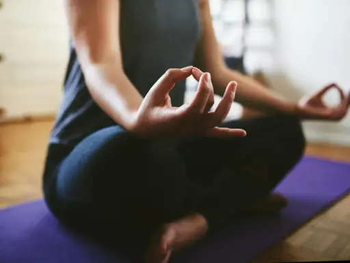 En person sitter i meditationsställning på en lila matta. Händerna vilar i knät med tumme och pekfinger sammanfogade, en vanlig gest för att visa hur man mediterar.