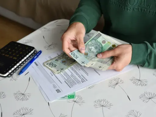 Ręce liczą banknoty 100 zł nad dokumentami ZUS. Nowe zasady zasiłków przedemerytalnych w praktyce.