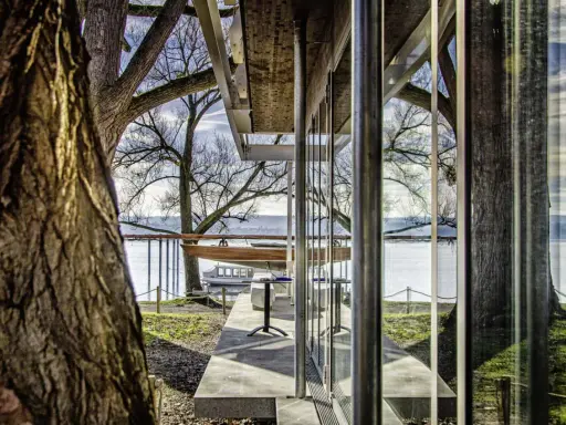 Ein Boot liegt aufgebockt am Ufer, daneben ein Tisch und Stühle. Die moderne Architektur des **Keller Werft**-Gebäudes spiegelt sich im Wasser.