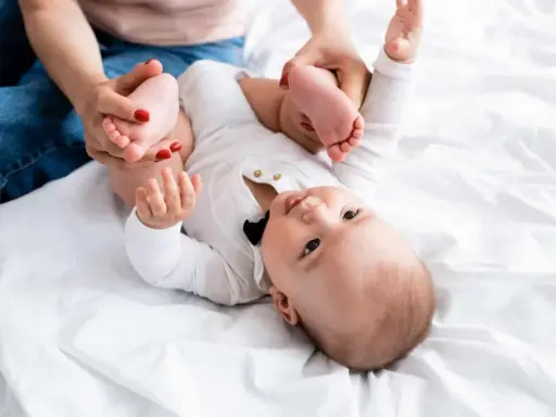Mama delikatnie ćwiczy nóżki niemowlaka, pomagając mu przy wzmożonym napięciu mięśniowym. Dziecko leży na białym tle.