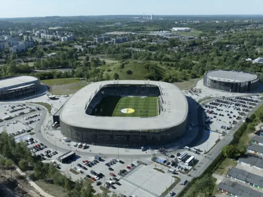 Widok z lotu ptaka na nowoczesny kompleks sportowy, w tym stadion piłkarski i dwie hale widowiskowe. To arena Sosnowiec, otoczona zielenią i parkingami.