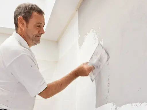 Mann verputzt eine Wand mit einer Kelle. Die Arbeit im Bad ist fast fertig.