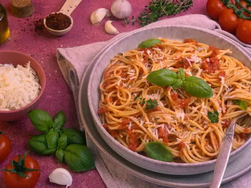 Ein Teller Spaghetti Napoli mit Tomatensauce, Basilikum und Parmesan. Daneben Zutaten wie Tomaten, Knoblauch und Olivenöl.