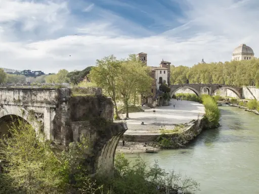 Un ponte rotto, con archi antichi e vegetazione rigogliosa, domina un fiume serpeggiante a Roma.