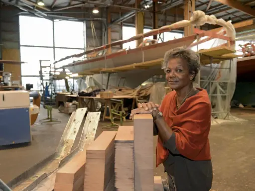 Jacqueline Tabarly, souriante, dans un atelier de construction navale, devant des blocs de bois et un voilier en cours de fabrication.