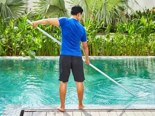 Hombre con camiseta azul y pantalones cortos negros limpia piscina de sal con un limpiafondos.