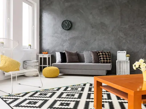 Nowoczesny salon z szarą ścianą, na której widać ciekawe efekty. Kanapa, biały fotel i geometryczny dywan tworzą przytulną przestrzeń.