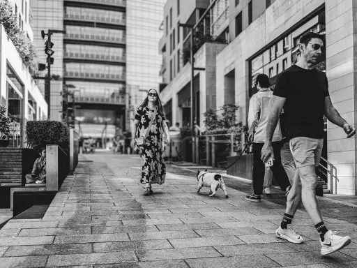 Street fotografia in bianco e nero: una donna con un vestito floreale passeggia con un cane, mentre altri camminano sullo sfondo.