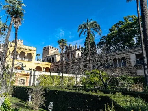 Sonniger Tag im Alcázar von Sevilla. Palmen ragen über kunstvoll verzierte Gebäude und gepflegte Gärten.
