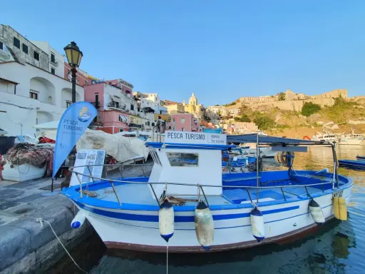 Barca bianca e blu ormeggiata, con bandiera "Pesca Turismo Isola di Procida". Case colorate sullo sfondo.