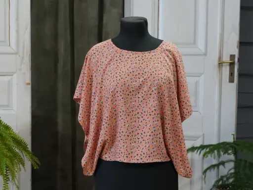 En persikofärgad blus med ett litet mönster av mörka prickar och små blommor. Blusen har en vid passform och korta, fladdermusliknande ärmar.