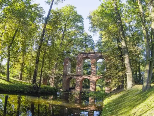 Kamienne ruiny mostu w Arkadii Nieborów, otoczone bujną zielenią i odbijające się w spokojnej wodzie.