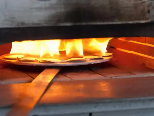 Des pièces de métal attendent d'être émaillées dans un four, sous des flammes vives. L'artisan émailleur surveille attentivement la cuisson.
