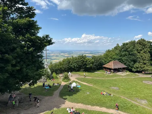 Ludzie odpoczywają na trawie i spacerują po szlakach na górze Ślęża. W oddali widać rozległą panoramę.