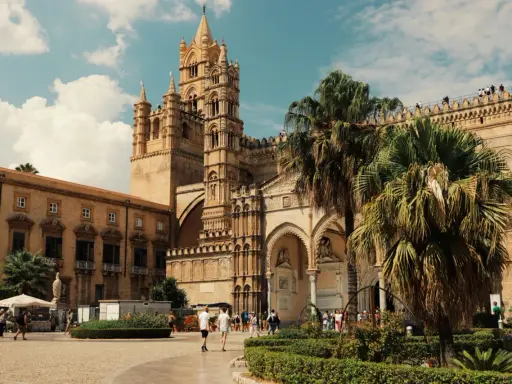 La Cattedrale di Palermo, un capolavoro di stili architettonici diversi, si erge maestosa sotto un cielo azzurro con palme rigogliose.