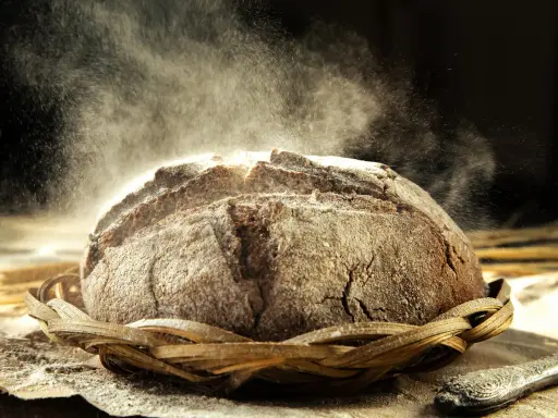 Un pain au seigle rustique, saupoudré de farine, repose dans un panier en osier. La poussière de farine danse dans la lumière, créant une atmosphère magique.