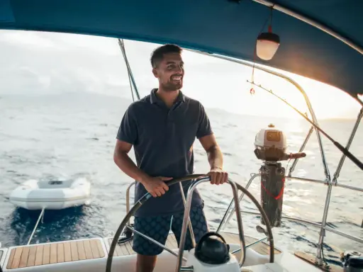 Un homme souriant est au gouvernail, prêt à barrer un bateau. Le soleil brille sur l'eau calme.