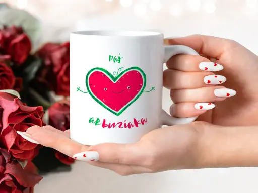 Biały kubek z zabawnym sercem-arbuzem i napisem "Daj buziaka", trzymany w dłoniach z paznokciami w serduszka, na tle czerwonych róż.