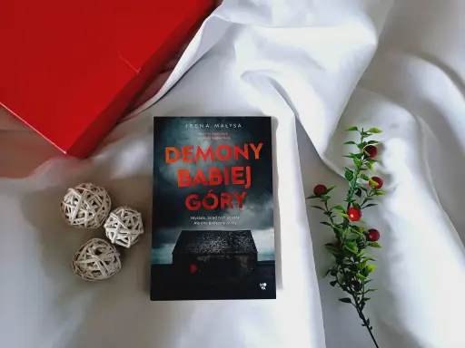 Książka "Demony Babiej Góry" Ireny Małysy, leżąca na białym jedwabiu, obok czerwonego pudełka i ozdobnych kulek. Historia, która zabiera daleko od Babiej Góry.