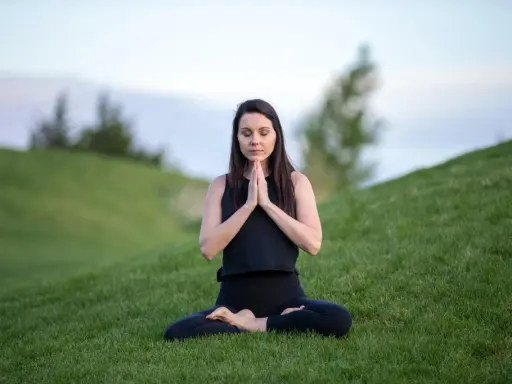 Kvinna i meditationsposition på en grön kulle, med slutna ögon och händerna i bön.