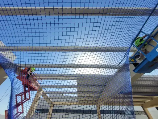 Trabajadores en plataformas elevadoras instalan una red de protección colectiva sobre una estructura en construcción, garantizando la seguridad.