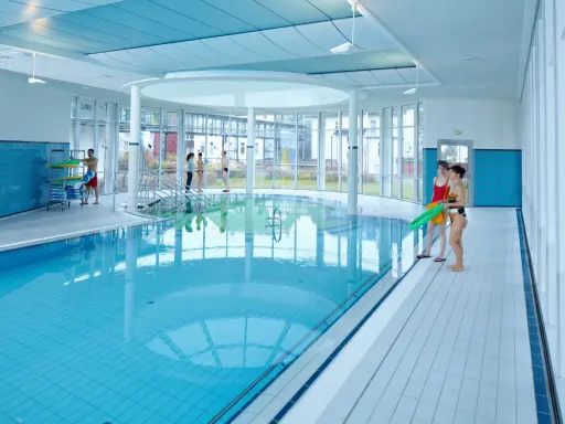 Menschen in Badekleidung am Beckenrand eines Hallenbades mit türkisfarbenem Wasser.