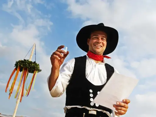 Ein Mann im Zimmermannsgewand hält ein Glas und eine Urkunde. Ein Richtkranz hängt am Himmel. Das ist ein Richtfest!