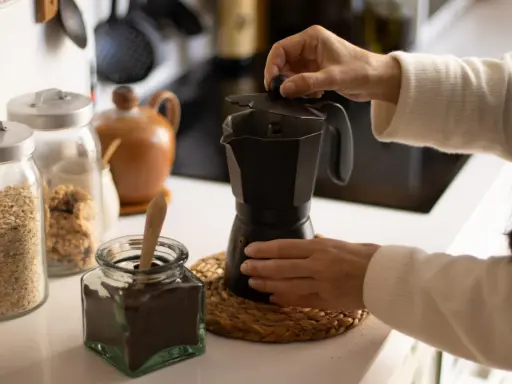 Manos preparando un café con alcohol en cafetera italiana. Hay frascos con avena y café molido.