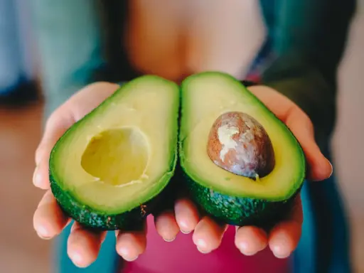 Zwei Hälften einer Avocado werden in Händen gehalten. Eine Hälfte hat den Kern, die andere ist leer. Perfekt, um die Avocado reifen zu lassen.