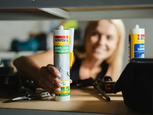 Kobieta trzyma tubkę akrylu lekkiego szpachlowego Soudal. Po jakim czasie można malować akryl? Po wyschnięciu i zeszlifowaniu.