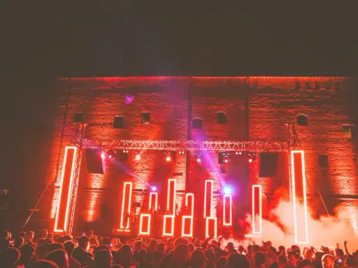 Folla in delirio al **mood festival**, illuminata da luci al neon rosse e fumo denso. La scena è un'esplosione di energia.