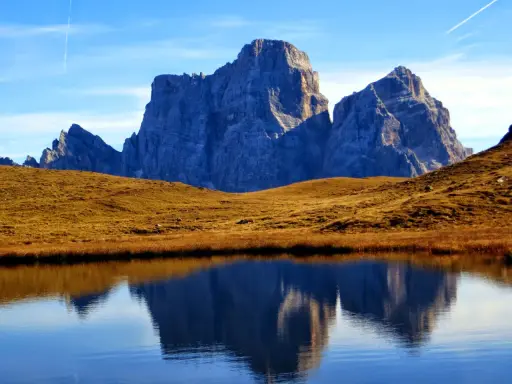 Riflessi maestosi delle Dolomiti nel lago delle Baste, con un prato dorato che separa le acque dalle vette imponenti.