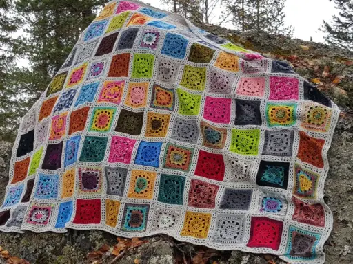 Ett färgglatt, virkat lapptäcke med många olika mönster och färger ligger utspritt över en mossig sten i skogen.